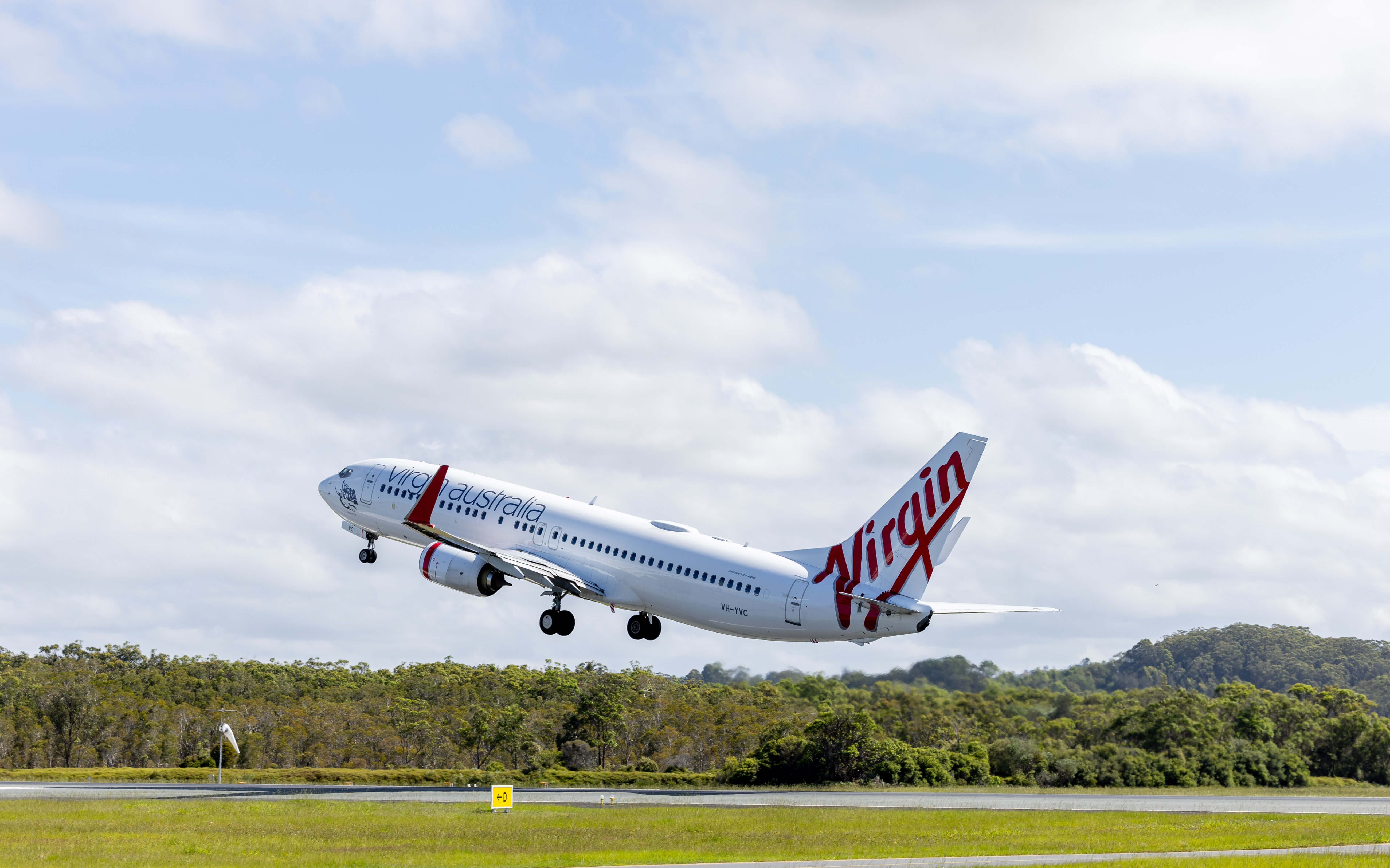 A Virgin Australia airplane, a Boeing 737, is captured mid-takeoff, ascending from the runway. The aircraft's white fuselage features the airline's red branding, with "Virgin" prominently displayed on the tail. The background consists of a partly cloudy sky and lush green trees near the airport. The landing gear is retracting as the plane gains altitude.