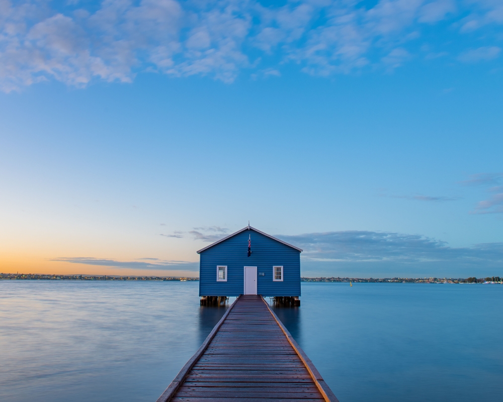 The iconic blue Crawley Edge Boatshed in Perth, Western Australia, sits at the end of a wooden jetty on the calm waters of the Swan River at sunrise, under a clear blue sky.
