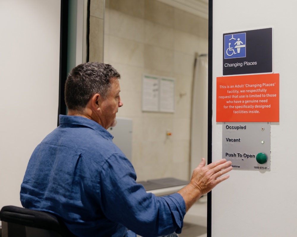 A man in a wheelchair reaches to press the button outside a Changing Places accessible facility. Signage on the door provides entry instructions and highlights accessibility features.
