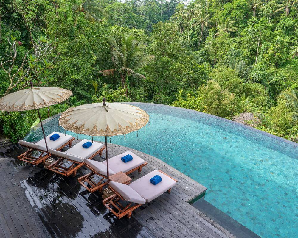 A tranquil infinity pool overlooking lush jungle in Bali, with four sun loungers topped with rolled blue towels and shaded by traditional Balinese umbrellas on a wooden deck.