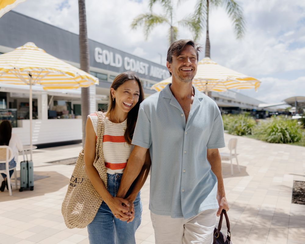A couple smiles while walking hand-in-hand outside Gold Coast Airport, passing yellow umbrellas and palm trees on a sunny day.