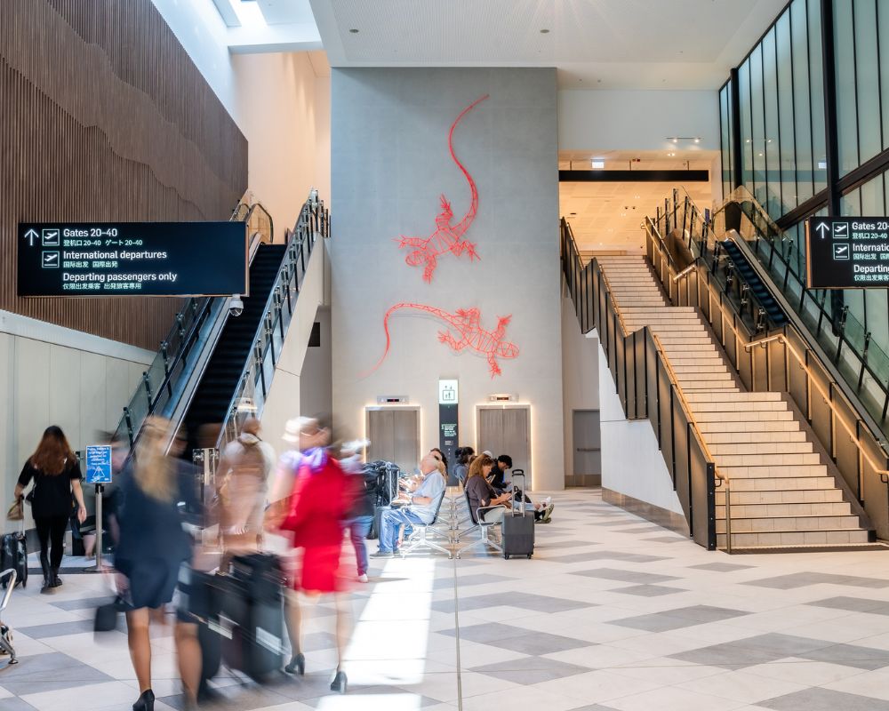 Passengers move through a modern airport terminal with escalators, a staircase, and red gecko-inspired wall art above a lift.