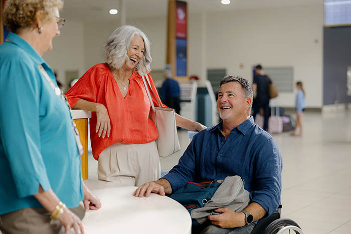 A man in a wheelchair smiles while speaking with two women at an airport assistance desk. One woman wears a red blouse and the other is in a teal uniform. The terminal background shows check-in kiosks and travellers with luggage.