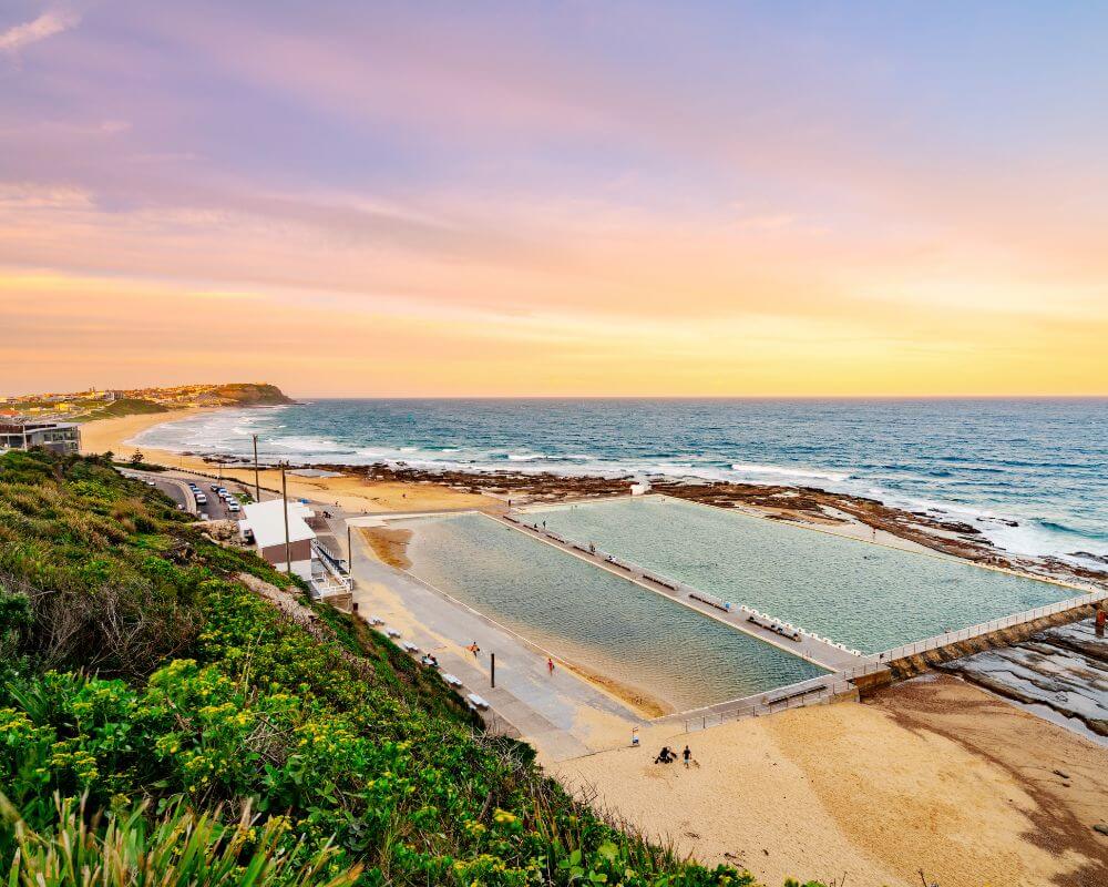 Coastal view of Newcastle Ocean Baths in New South Wales at sunset, with swimmers enjoying the tidal pool beside the sandy beach and waves rolling in from the ocean under a pastel-coloured sky.
