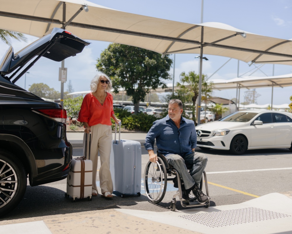A man in a wheelchair and a woman with luggage stand next to an open car boot at an airport drop-off zone. The area is covered by shade sails, with other vehicles and palm trees in the background.