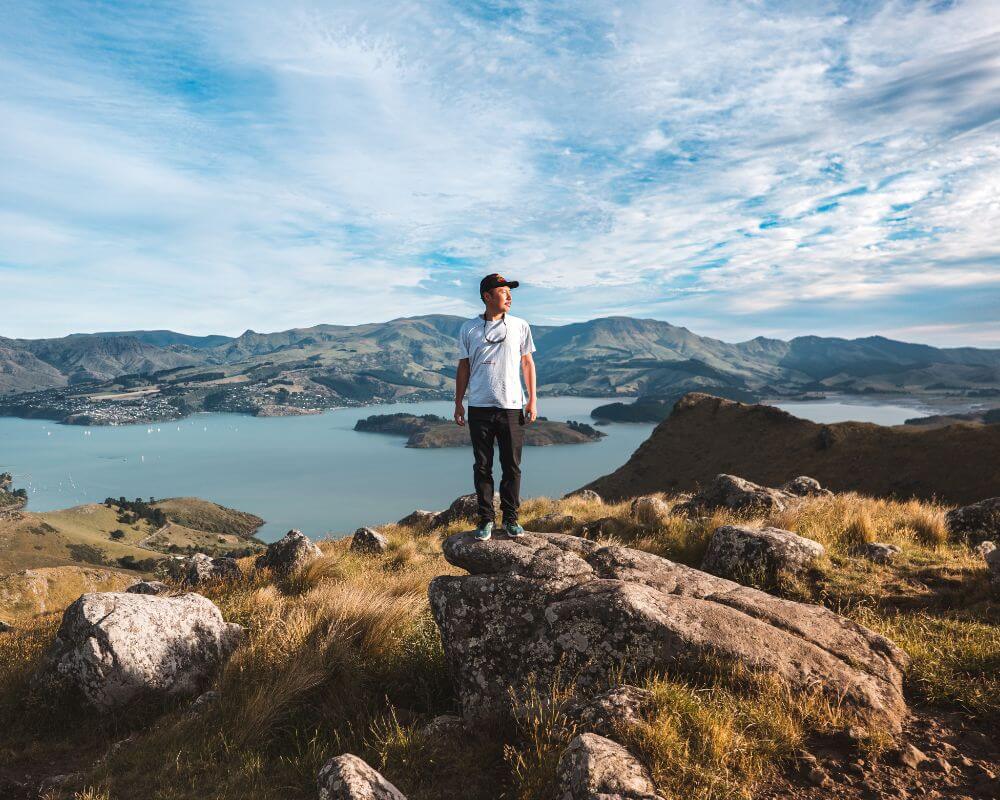 A man stands on a rocky hilltop with panoramic views of Christchurch’s Lyttelton Harbour in New Zealand. Surrounded by golden grass and rugged terrain, he overlooks the deep blue bay and rolling hills under a dynamic sky.
