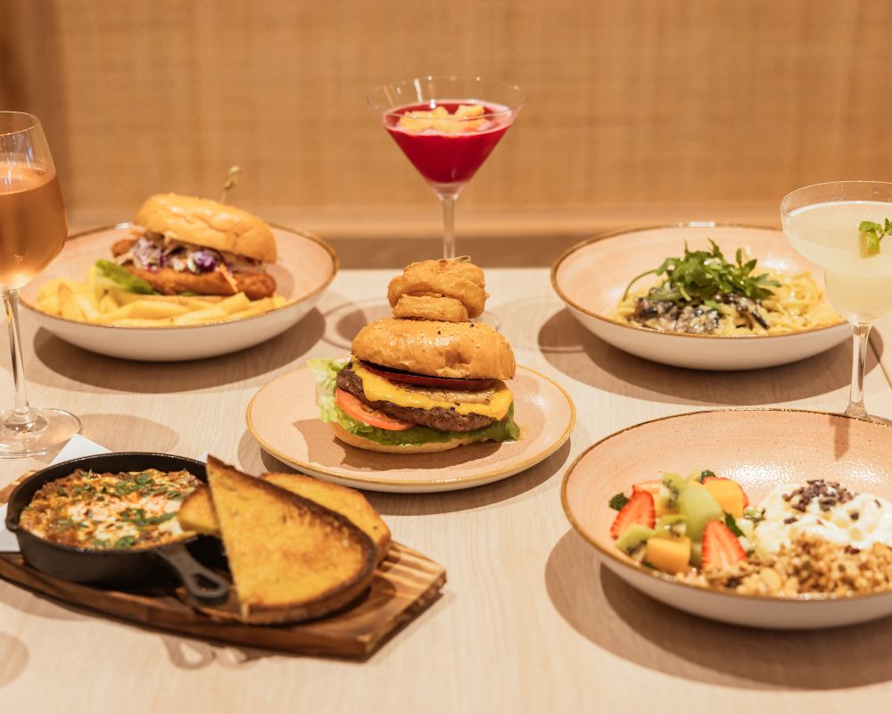 A table spread of gourmet dishes including burgers, pasta, bread with dips, a fresh fruit bowl, and cocktails, set in a stylish dining environment.