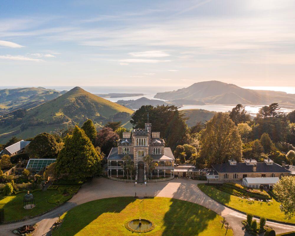 Aerial view of Larnach Castle near Dunedin, New Zealand, surrounded by lush gardens and rolling green hills. In the distance, Otago Harbour glistens under the soft light of late afternoon.