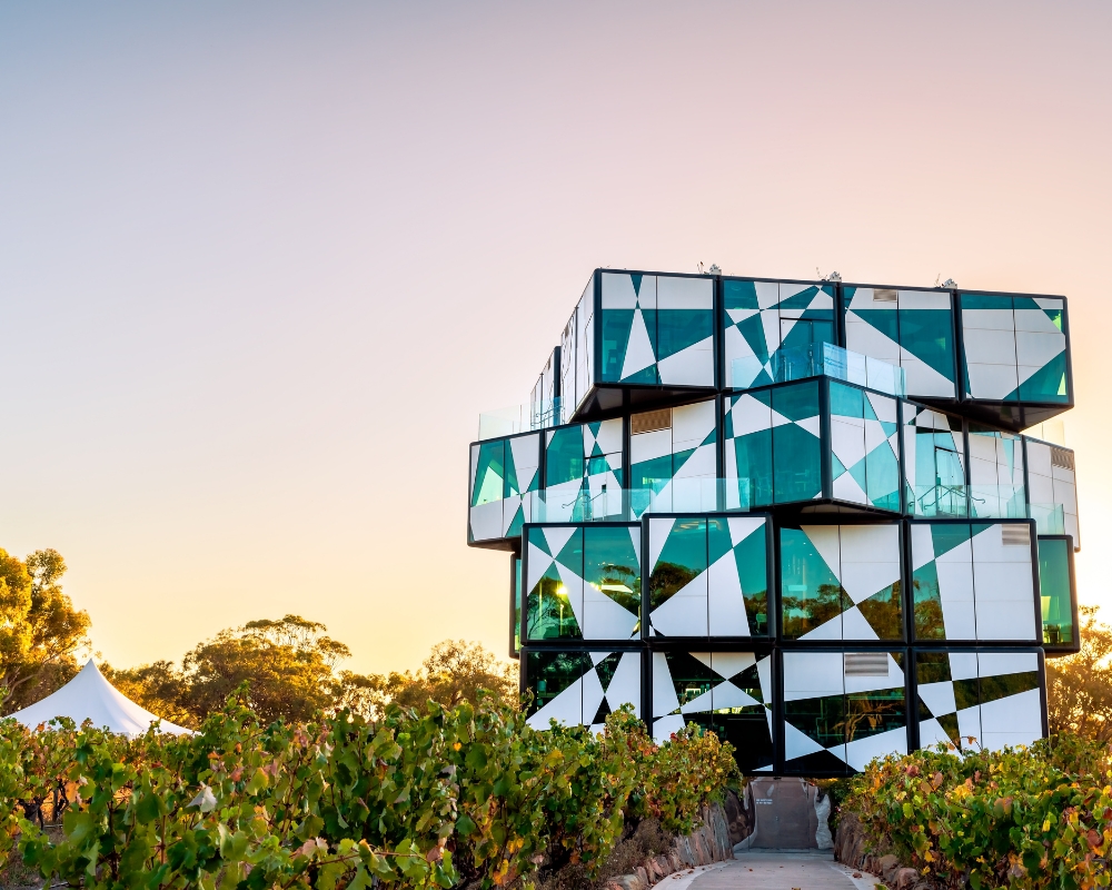 The d’Arenberg Cube in McLaren Vale, South Australia, a striking multi-storey building with an abstract, cube-like design made of glass panels in geometric patterns, surrounded by vineyards at sunset.