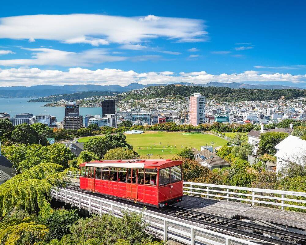 Iconic red Wellington Cable Car travelling uphill with panoramic views over Wellington city, lush gardens, and the harbour on a clear day.