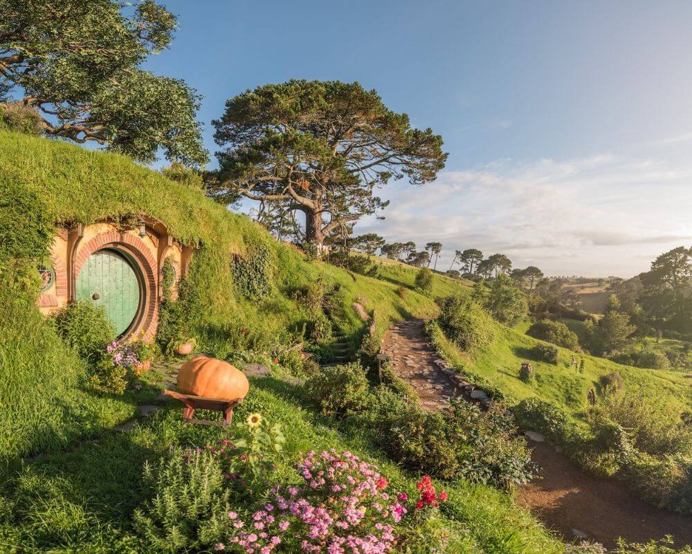 A picturesque view of Hobbiton in Matamata, New Zealand, featuring a round green hobbit door nestled in a hillside, surrounded by flowers and a winding garden path under golden morning light.