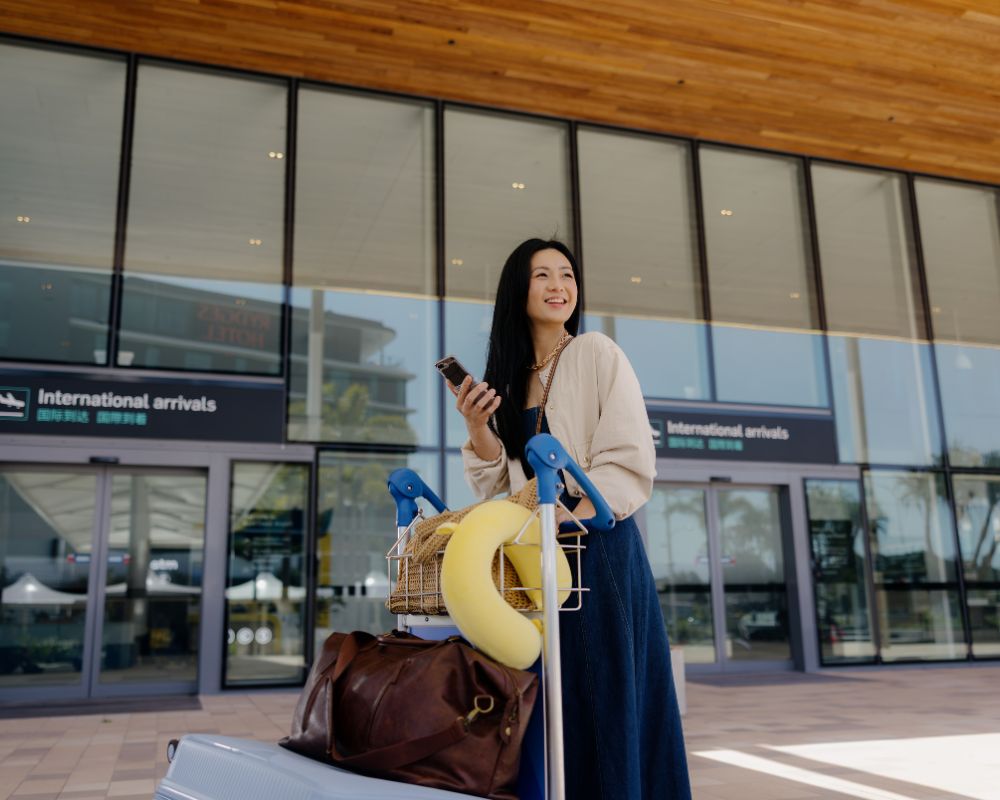 A woman stands outside the International Arrivals entrance at Gold Coast Airport with a luggage trolley, holding her phone and smiling.