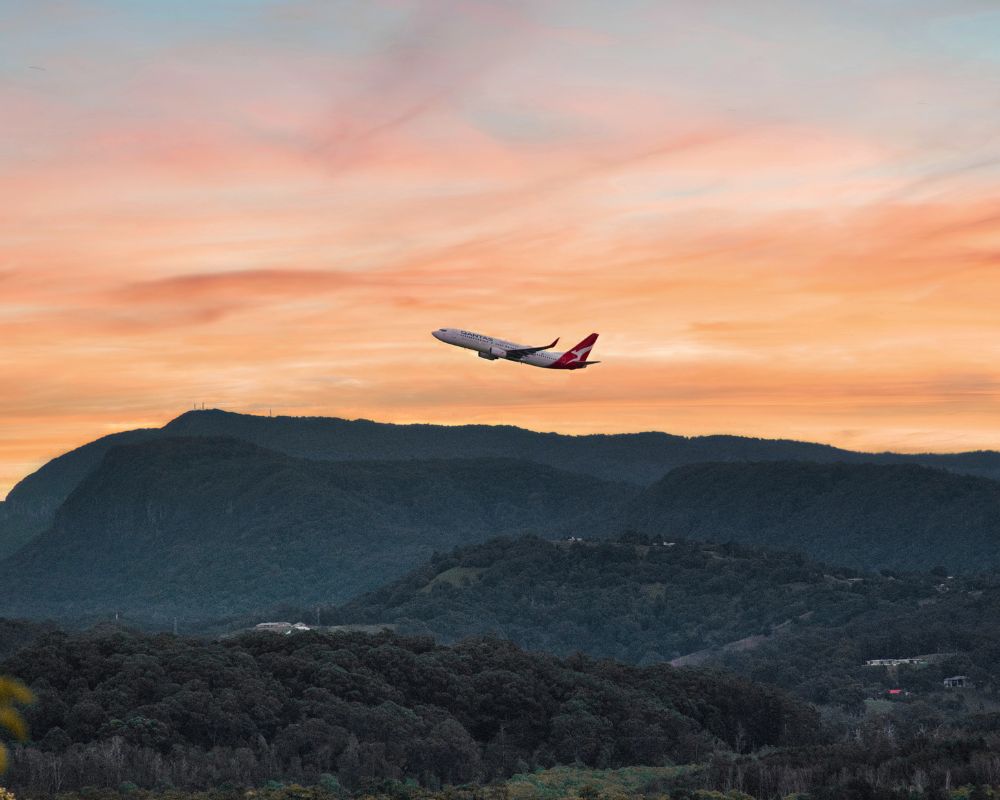 A Qantas aircraft takes off into a vibrant orange and pink sunset sky, soaring above lush green hills and forested mountains near Gold Coast Airport.
