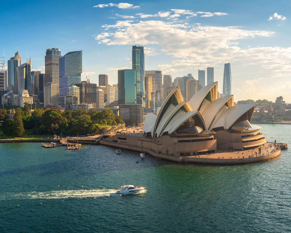 Aerial view of the Sydney Opera House at sunset, surrounded by Sydney Harbour and the city skyline. A white boat cruises across the sparkling water in the foreground.